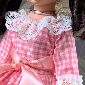 Puede incluir: Un vestido de cuadros vichy rosa y blanco con un cuello de encaje blanco y un lazo de sat&eacute;n rosa. El vestido tiene mangas largas y un dobladillo con volantes.