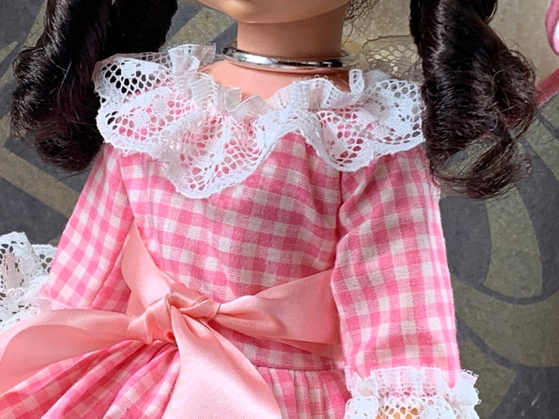 Puede incluir: Un vestido de cuadros vichy rosa y blanco con un cuello de encaje blanco y un lazo de sat&eacute;n rosa. El vestido tiene mangas largas y un dobladillo con volantes.