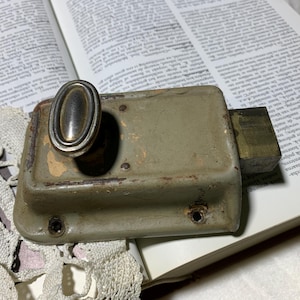 Op de afbeelding: Een antieke deursluiting met een aangetaste zilveren ovale handgreep en een rechthoekig olijfgroen lichaam. De sluiting ligt op een open boek met vervaagde tekst en kant.