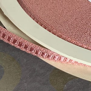 May include: Close-up of a spool of pink ribbon with a textured surface. The ribbon is a muted rose color and has a woven design. The spool is white and the background is a patterned surface.