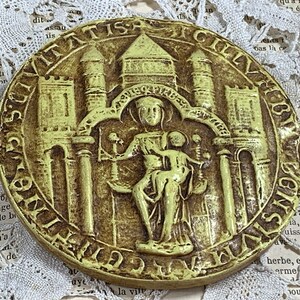 Può includere: Un sigillo metallico circolare, di colore verde, in rilievo con il testo "CIVITATIS" e "SIGILLVM" attorno al bordo. Al centro del sigillo, una scultura in rilievo di una donna che tiene un bambino, entrambi seduti su un trono. La donna indossa una corona e il bambino tiene uno scettro.