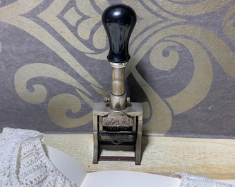 May include: Vintage metal date stamp with a black handle. The stamp has a silver metal body and a rectangular base. The background features a decorative pattern in gold and gray.