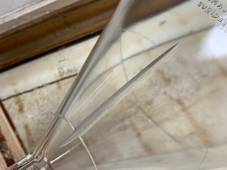 May include: A clear glass funnel with a long, thin, metal rod inside. The funnel is resting on a white surface.