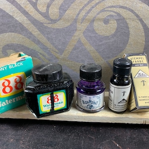 May include: Four bottles of ink, two black and two purple, with labels that read "88 Waterman", "Bon Point", and "T". The bottles are sitting on a wooden surface. A box of ink labeled "Ebony Black 88 Waterman" is in the background.