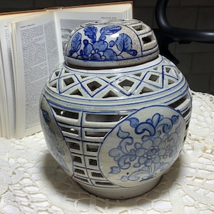 Op de afbeelding: Blauw-witte keramische pot met deksel, versierd met bloemmotieven. De pot heeft een opengewerkt roosterpatroon op de body. Een open boek is zichtbaar op de achtergrond, naast de pot.