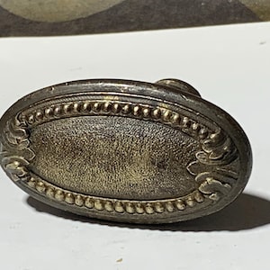 May include: An antique oval door knob with an aged, tarnished brass finish. The knob features a detailed beaded border and ornate floral-like designs on each end. The center has a textured surface.