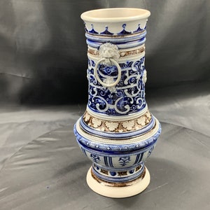 Op de afbeelding: Een blauwe en witte keramische vaas met ingewikkelde bloem- en geometrische patronen. De vaas heeft een ringvormige handgreep met een leeuwenkop aan elke kant. De vaas is versierd met bruine accenten.