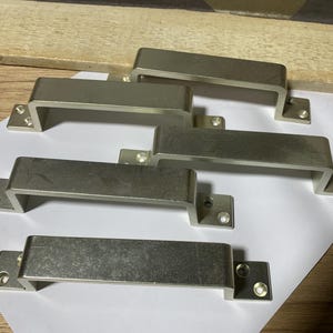 May include: Five brushed nickel cabinet pulls arranged on a white surface. Each pull has a rectangular bar and two mounting plates with screw holes. The pulls are a neutral silver color.