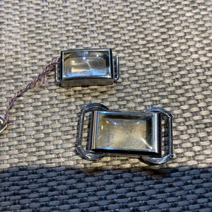 Op de afbeelding: Twee rechthoekige horlogegespen met een zilverkleurig metalen frame en een helder, glasachtig midden. Eén gesp heeft een label met het woord "ARSA" erop. De gespen liggen op een getextureerd, grijs oppervlak.