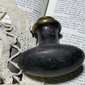 May include: A dark, oval-shaped door knob with a brass collar. The knob has a matte, black finish with signs of wear. The background includes a vintage book and lace.