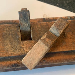 May include: A vintage wooden hand plane featuring a dark brown, weathered wood body with a rectangular opening. A metallic, oxidized blade is inserted vertically into the slot, while a separate, smaller wooden wedge piece rests diagonally across.