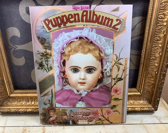May include: A book titled "Puppen Album 2" featuring a vintage porcelain doll with a pink dress and a white lace bonnet. The doll has blue eyes and a delicate facial expression. The book cover also includes a floral design and the text "Französische Porzellanpuppen" in German.