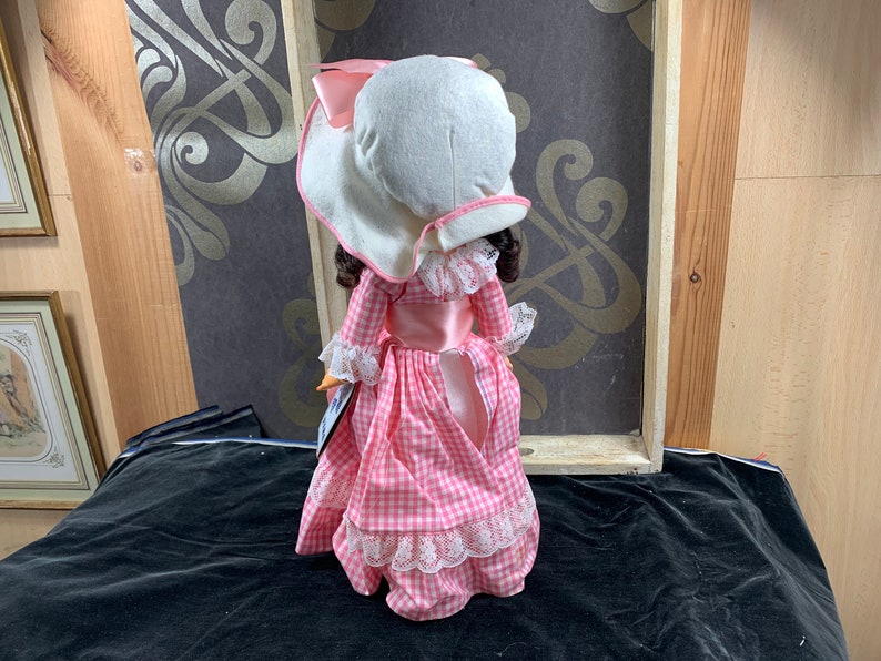 Puede incluir: Una mu&ntilde;eca de porcelana que lleva un vestido de cuadros vichy rosa y blanco con un ribete de encaje blanco. La mu&ntilde;eca lleva un gorro blanco con una cinta rosa.