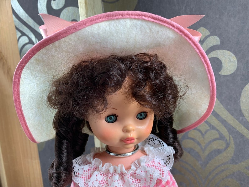 Puede incluir: Una mu&ntilde;eca vintage con el pelo rizado marr&oacute;n, ojos azules y un sombrero de sol rosa y blanco. La mu&ntilde;eca lleva un vestido de cuadros rosa y blanco con ribete de encaje blanco.