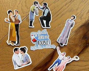 Peut inclure: Une collection d'autocollants avec des illustrations de personnages en costumes d'époque. Les autocollants représentent des scènes de personnes interagissant, dont un couple dansant et un homme agenouillé devant un enfant. Un autocollant porte le texte "My friends you've got trouble! Right here in river city."