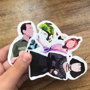 May include: A collection of die-cut stickers featuring stylized illustrations of characters. The stickers depict figures in various colors, including green, black, pink, and blue. The stickers are held against a wooden background.
