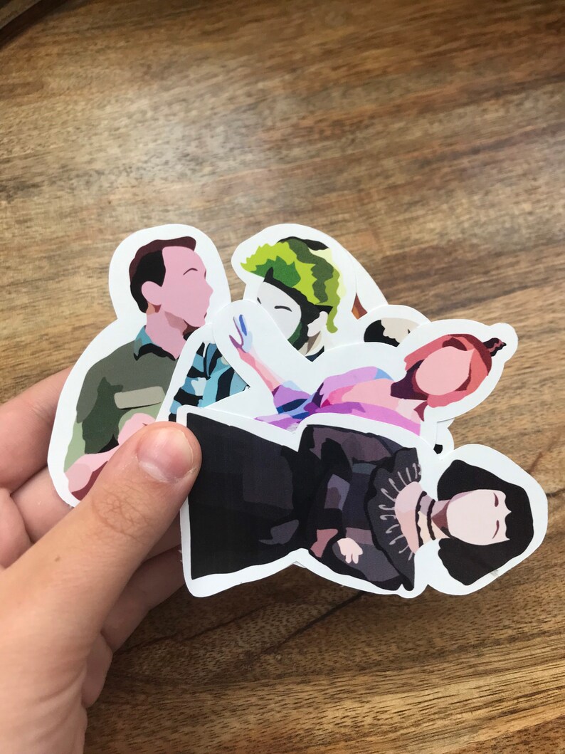 May include: A collection of die-cut stickers featuring stylized illustrations of characters. The stickers depict figures in various colors, including green, black, pink, and blue. The stickers are held against a wooden background.