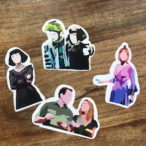 May include: A collection of five die-cut stickers featuring stylized illustrations of characters. The stickers depict figures in various outfits, with a focus on bold colors and simple shapes. The stickers are on a wooden surface.