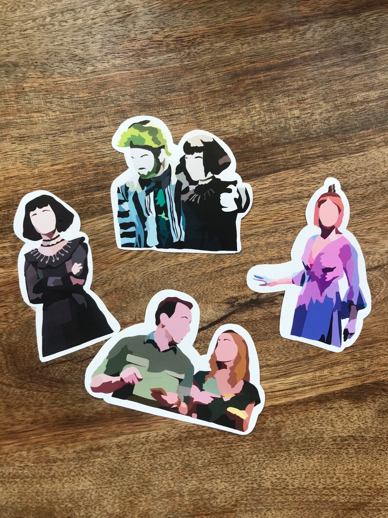 May include: A collection of five die-cut stickers featuring stylized illustrations of characters. The stickers depict figures in various outfits, with a focus on bold colors and simple shapes. The stickers are on a wooden surface.