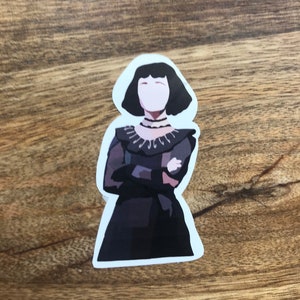 May include: A sticker featuring a stylized illustration of a woman in a dark dress with a high collar and short black hair. The woman has her arms crossed and is set against a white background. The sticker is likely for decorative purposes.