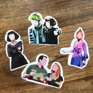 May include: A collection of five die-cut stickers featuring stylized illustrations of characters. The stickers depict figures in various outfits, with a range of colors including black, green, blue, and pink. The stickers are on a wooden surface.