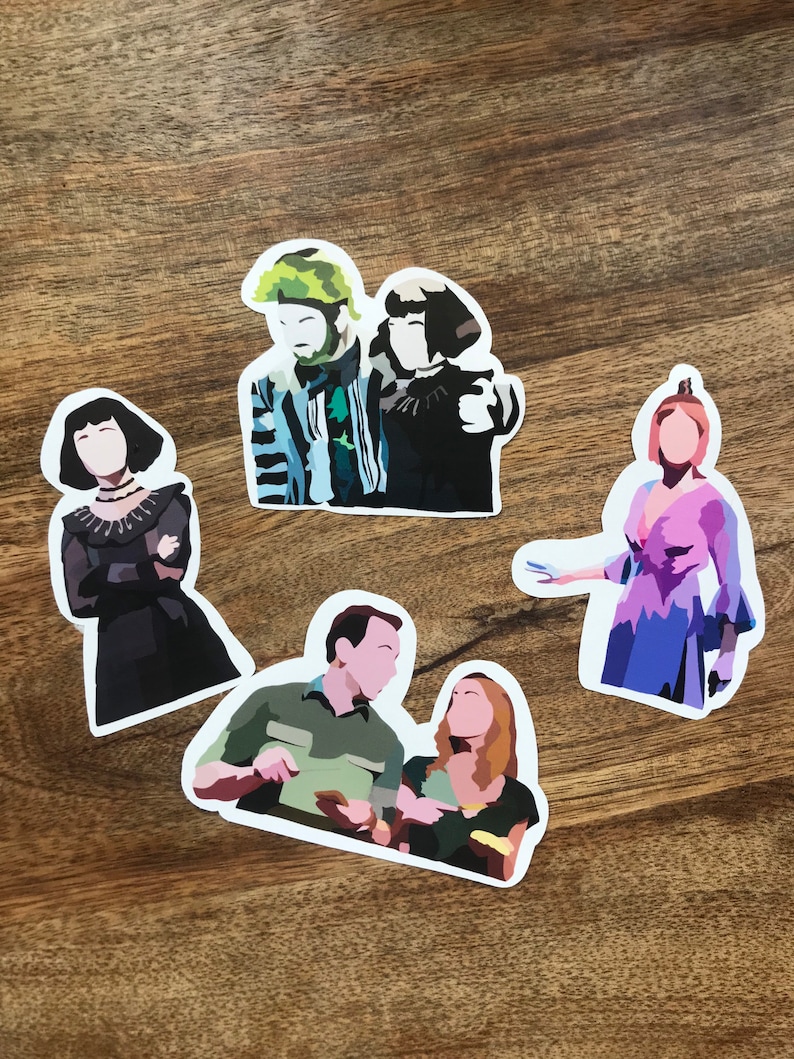 May include: A collection of five die-cut stickers featuring stylized illustrations of characters. The stickers depict figures in various outfits, with a range of colors including black, green, blue, and pink. The stickers are on a wooden surface.