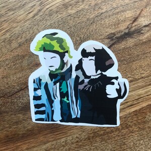 May include: A sticker featuring a stylized illustration of two characters. One character has a green and brown camouflage-patterned hair and beard, and the other has short black hair. The first character is wearing a striped suit with blue and black stripes.