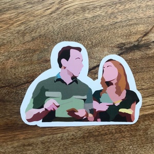 May include: A die-cut sticker featuring a stylized illustration of two people. The man is wearing a green shirt, and the woman is wearing a dark green top. The sticker has a white border and is set against a wooden background.