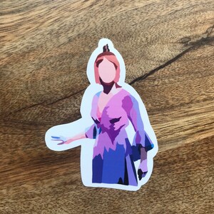 May include: A die-cut sticker featuring a stylized illustration of a person in a purple and blue dress. The sticker has a white border and is placed on a wooden surface. The person's hair is a light reddish-brown color.