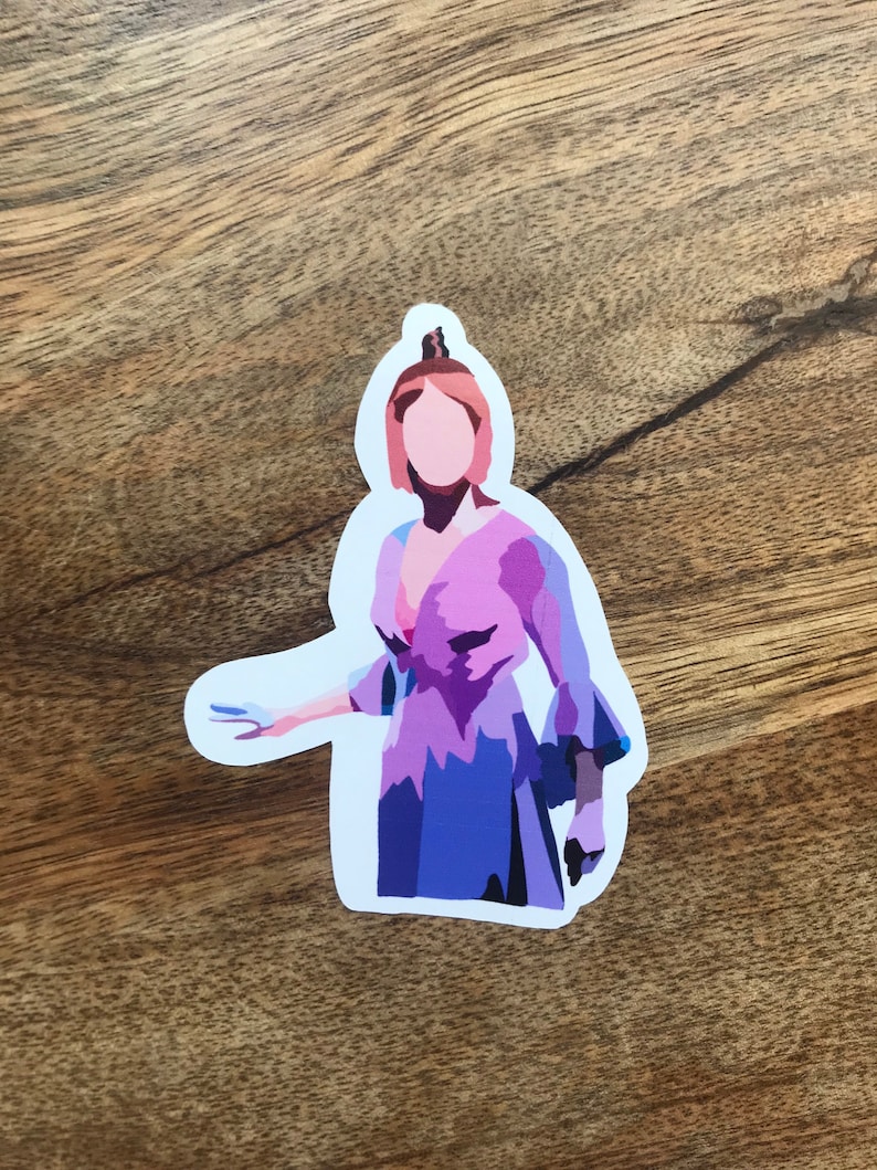 May include: A die-cut sticker featuring a stylized illustration of a person in a purple and blue dress. The sticker has a white border and is placed on a wooden surface. The person's hair is a light reddish-brown color.