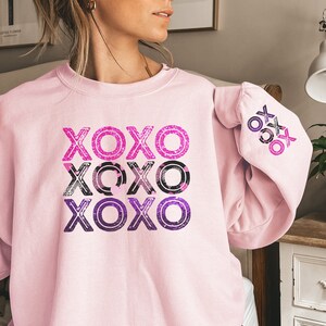 May include: A pink sweatshirt with a graphic print of the word "XOXO" repeated three times in a gradient of pink, purple, and black. The sweatshirt has a relaxed fit and long sleeves.