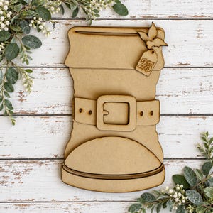 May include: A light brown wooden cutout shaped like a boot with a cuff at the top, a belt with a square buckle across the middle, and a small tag attached to the side featuring the number 25 and a holly leaf design on top.