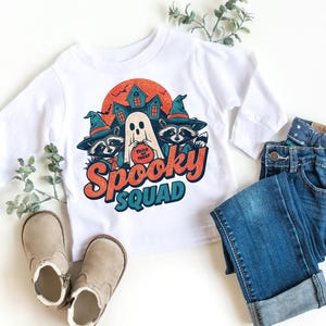 Könnte beinhalten: Weißes Langarmshirt mit einem Halloween-Motiv. Das Motiv zeigt einen Geist, Waschbären mit Hexenhüten und den Schriftzug "Spooky Squad". Das Outfit wird durch blaue Jeans und beige Schuhe ergänzt, ideal für Halloween.