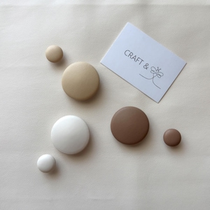 Peut inclure: Assortiment de boutons ronds en bois décoratifs de couleur beige, marron et blanc. Les boutons sont disposés sur une surface blanche avec une carte de visite sur laquelle est inscrit "CRAFT &" avec un motif de fleur stylisé.