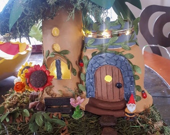 May include: A miniature fairy house with a green leafy roof, a brown door, and a small gnome sitting on the steps. The house is surrounded by moss and greenery, and there is a small swing set in the foreground.
