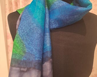May include: A blue, green, and gray silk scarf with a watercolor-like design. The scarf is folded and draped over a mannequin.