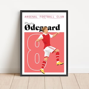 May include: A framed poster featuring a stylized illustration of Martin Ødegaard in an Arsenal football kit. The poster has a red background with a large number 8. The text "ARSENAL FOOTBALL CLUB" is at the top.