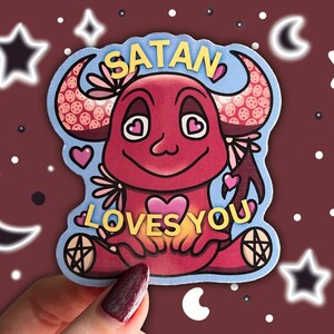 May include: A red cartoon devil with horns and a heart in its hands. The devil is surrounded by a blue background with pink flowers and hearts. The text "Satan Loves You" is written in yellow letters.