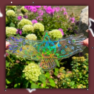 May include: A holographic sticker of a bird with outstretched wings. The bird has a pentagon star in its center with a circle around it. The sticker is iridescent and has a rainbow effect.