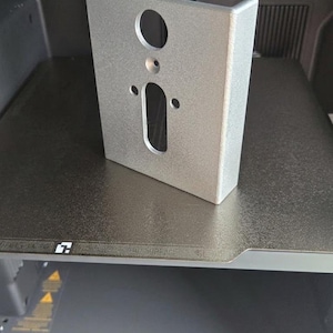 May include: A silver-colored, U-shaped metal bracket with a textured finish. The front face features a circular hole at the top, a smaller centered hole below it, and a vertical rectangular slot flanked by two smaller circular holes near the bottom.