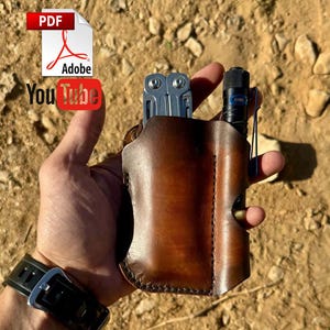 May include: A brown leather multi-tool sheath holding a silver multi-tool and a black flashlight. The sheath is hand-stitched and has a loop for a pen or small tool. The background is a dirt path.