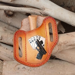May include: A brown leather holster with red stitching. The holster features a black handgun design and a playing card graphic with the 10, Jack, Queen, and King of Spades. The holster has a wooden peg.
