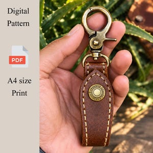 May include: A brown leather keychain featuring a brass-toned swivel lobster clasp and a central circular brass snap button with embossed text. The leather is stitched with light-colored thread along the edges. Text reads Digital Pattern PDF A4 size Print.