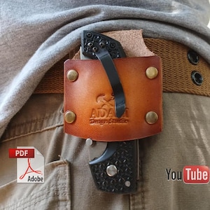 May include: A brown leather knife sheath with a black folding knife clipped to a belt. The sheath has the text "ADA Design Studio" and is secured with brass rivets. The knife is partially visible.