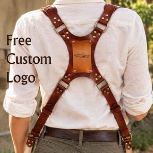 May include: A brown leather camera harness with brass hardware and a central back plate featuring the text Von Lawrence. The harness includes adjustable straps and metal clips attached to two cameras. The text Free Custom Logo is visible.