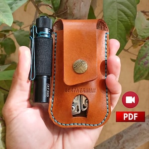 May include: A black flashlight with a blue clip beside a brown leather Leatherman sheath. The sheath features a metal button and blue stitching. The word "Leatherman" is embossed on the front.