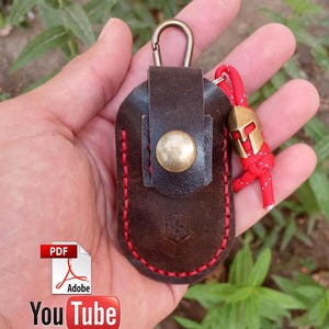 May include: A brown leather key chain with a metal clasp and a red cord with a metal charm. The key chain has a button on the front and is stitched with red thread.