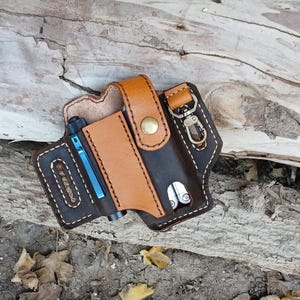 May include: A brown leather multi-tool holder with a tan leather flap and a metal snap closure. The holder has a small pocket for a pen or pencil and a larger pocket for a multi-tool. There is a metal key ring attached to the holder.