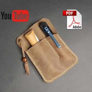 May include: A brown leather pouch with a flap and a stitched edge. The pouch has two pockets, one for a small knife and one for a pen. The pouch has a brown leather strap with a wooden bead.