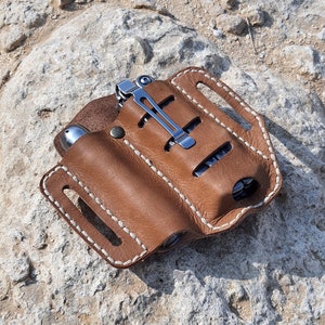 Op de afbeelding: Een bruine leren multitool-tas met witte stiksels. De tas heeft drie vakken voor gereedschap en een metalen clip.